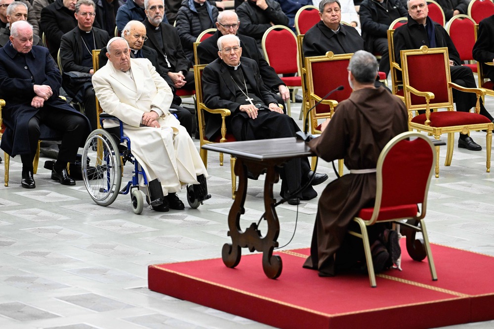 Capuchin Fr. Roberto Pasolini, Papal Preacher, Vatican City, Dec. 6, 2024. Pasolini sits on red dais, shown from behind, Facing his is Pope Francis in wheel chair, and many Cardinals clothed in black clerics and seated in red chairs.