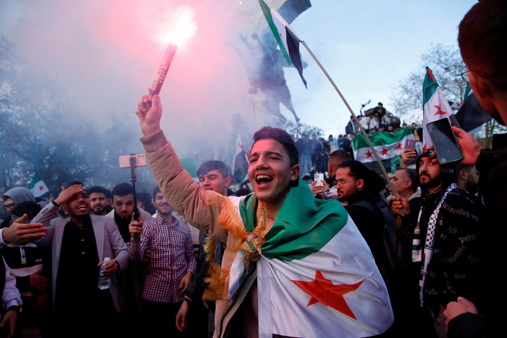 Syrian refugees in Turkey celebrate the fall of Assad regime, Istanbul, Dec. 8, 2024. Overjoyed young man wearing Syrian flag, holds flare and smiles; behind him is crowd.