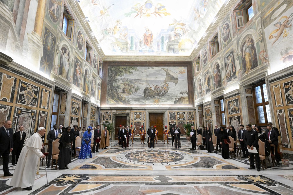 Pope Francis, receiving letters of new Diplomats, Vatican, Dec. 7, 2024. Large frescoed room; diplomats sit in u-shaped formation of chairs, Pope enters walking on cane.