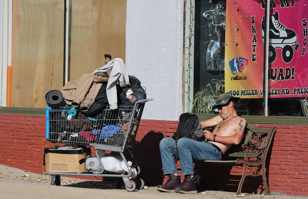 Homeless man, Prescott, Ariz., Dec. 9, 2024. Man sits shirtless on bench, next to him is shopping cart filled with posessions.