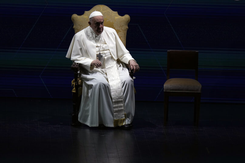 Pope Francis, Rome, May 10, 2024. Francis sits on chair, highlighted by light, surrounded by shadow.