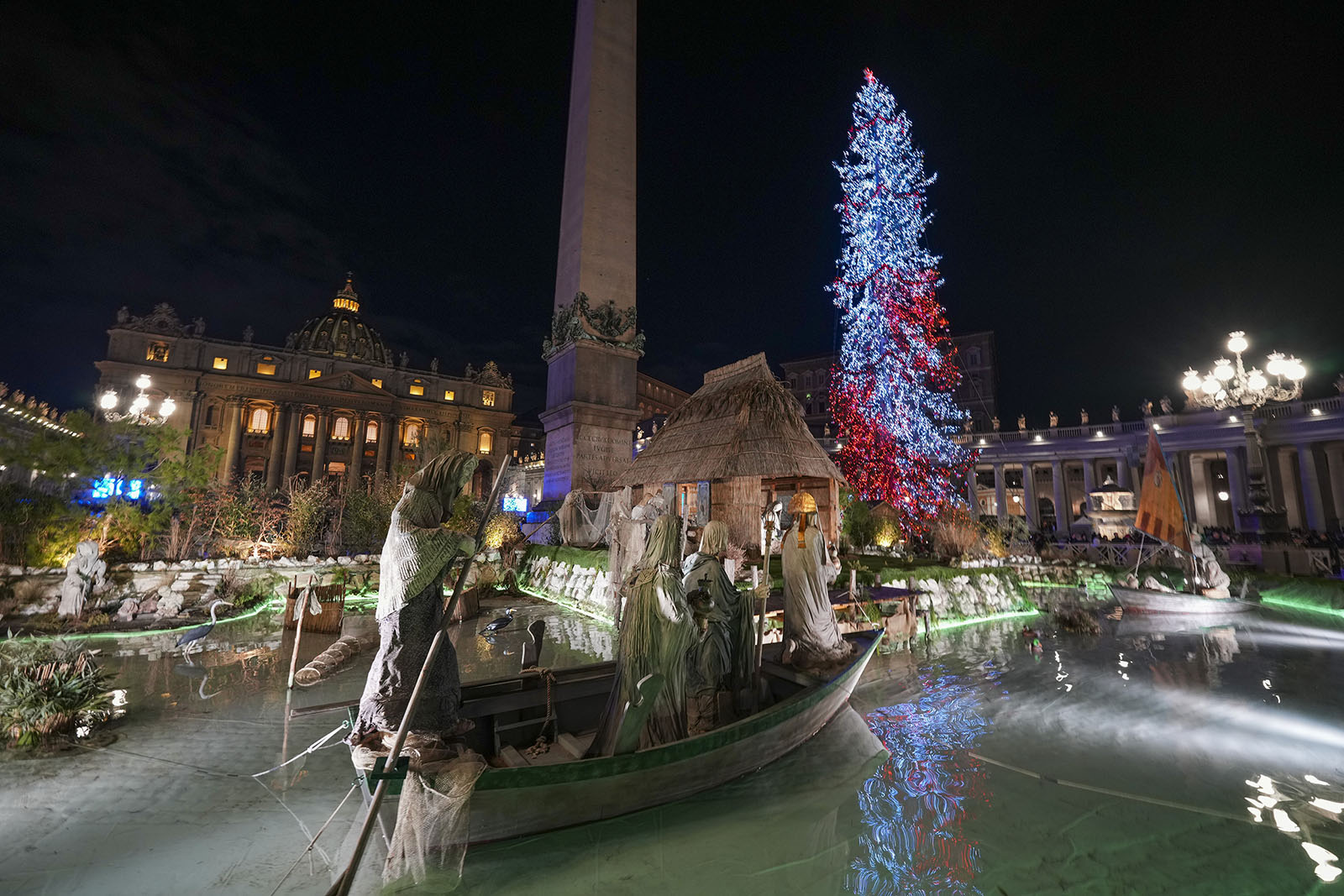 Vatican Christmas tree and nativity scene, 2024. Illuminated tree and Vatican obelisk visible in background; foregrounded is part of nativity scene.