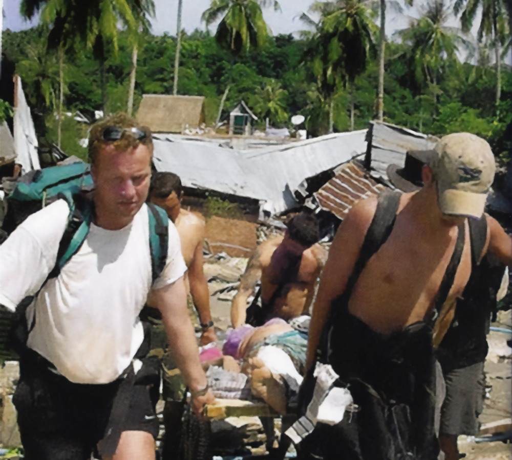 Deacon Ted Berghausen, Koh Phi Phi, Thailand, 2004. The deacon, shirtless with face obscured by bill of cap, carries a person with the help of others.