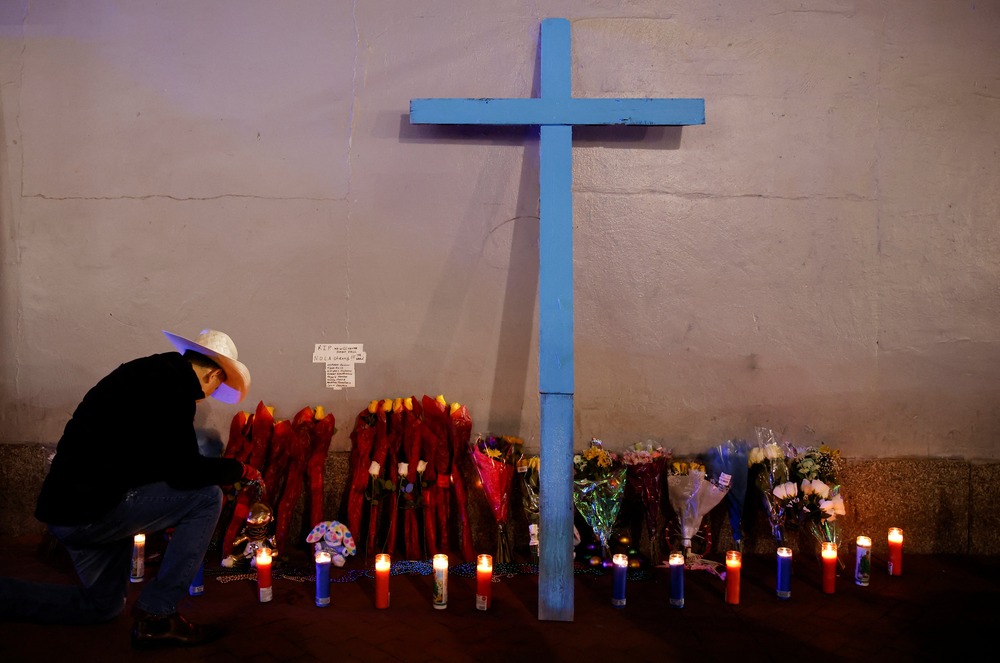 Makeshift memorial after Jan. 2025 French Quarter attack, New Orleans, Jan. 2, 2025. A cross leans against a wall lined with votive candles, on left a man kneels.