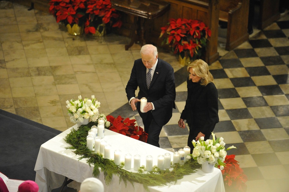 President Biden and First Lady Jill Biden, interfaith memorial for New Orleans attack victims, St. Louis Cathedral, New Orleans, Jan. 6, 2024. President Biden and First Lady Jill Biden wear black and approach altar adorned with greenery and candles. The president holds large white pillar candle.
