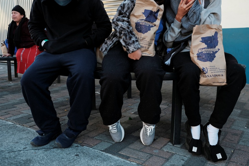 Migrants in Guatemala after U.S deportation flight to Guatemala City, Jan. 27, 2025. Three people with faces out of frame sit on a bench, two hold paper bags.