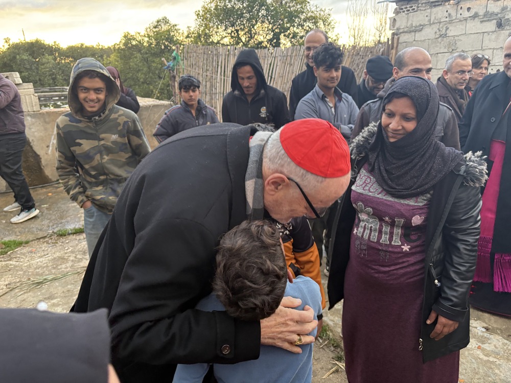 Cardinal Michael Czerny, Syrian refugees, Camp 004, Kfar Dlaqous, Lebanon, Feb. 21, 2025. Czerby bends down to guf two children, their mother watches joyfully; many others in background.