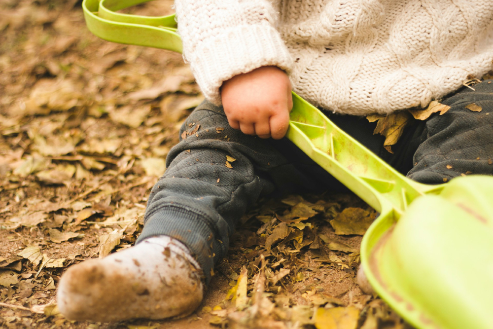 Child playing with dirt and leaves A cropped view of a young child wearing a white sweater and grey pants covered in dirt while holding a green plastic shovel and sitting in a pile of brown leaves.