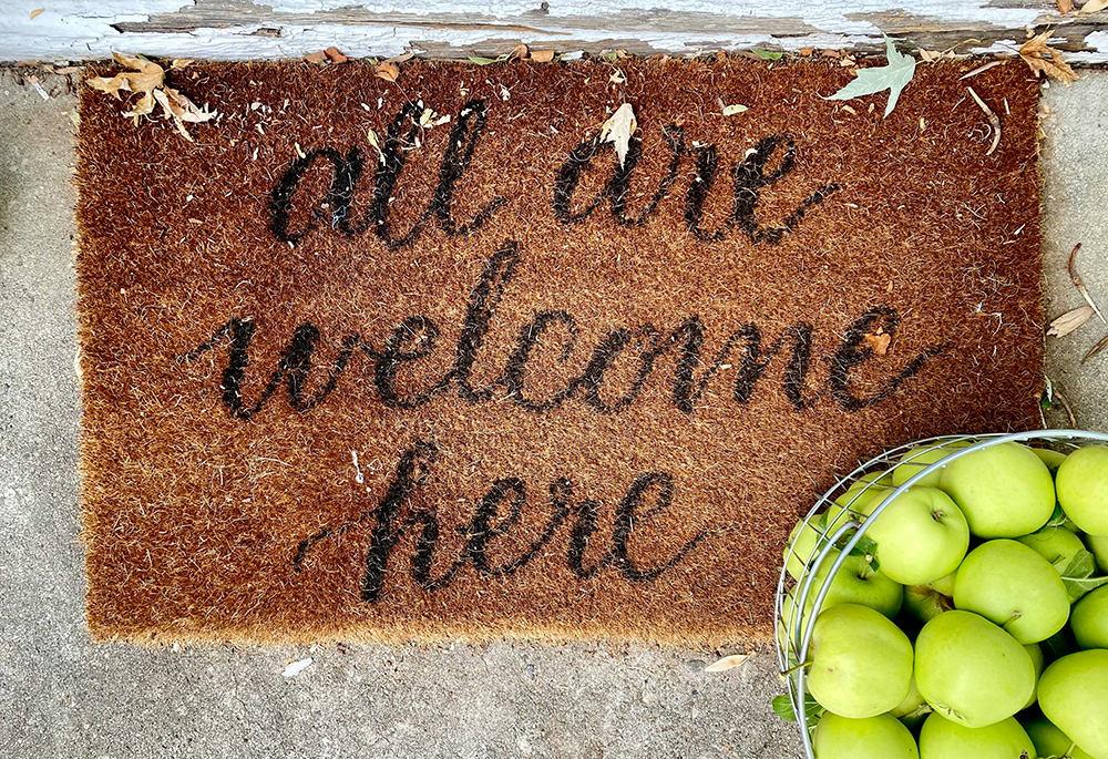 A brown welcome rug reads "all are welcome here" in black, cursive letting. The rug lies upon a front porch, with a container of green apples to its right. (Unsplash/LeeAnn Cline)