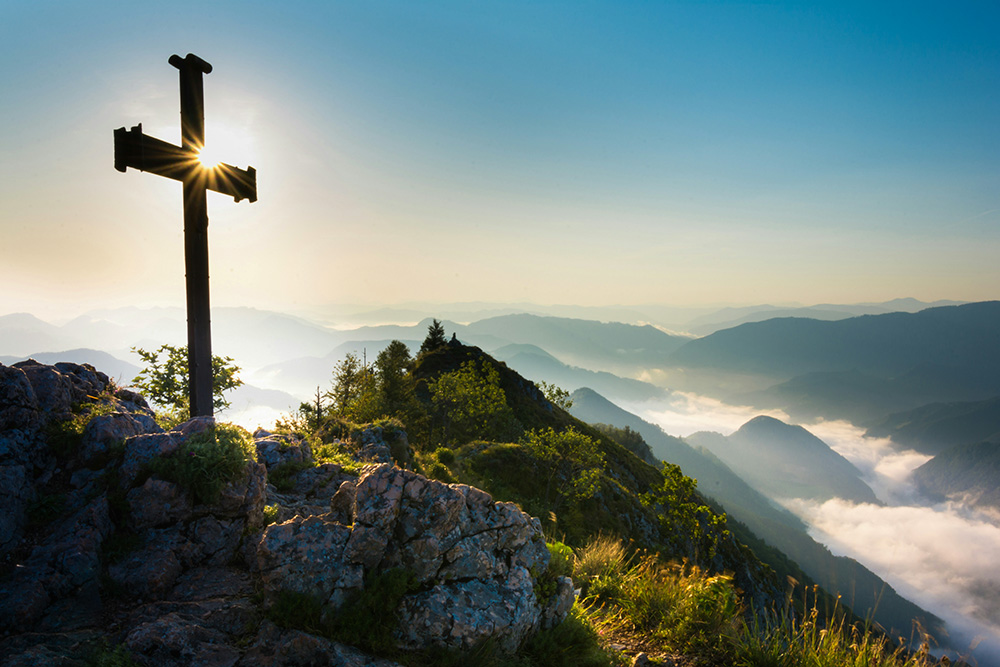 Cross lit by sunlight atop a mountain range (Unsplash/Theo Crazzolara)