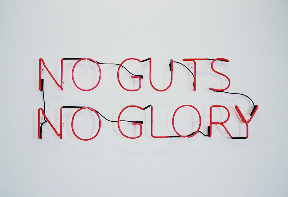 A red neon sign reads "No guts no glory" against a white wall. (Unsplash/Artem Bryzgalov)