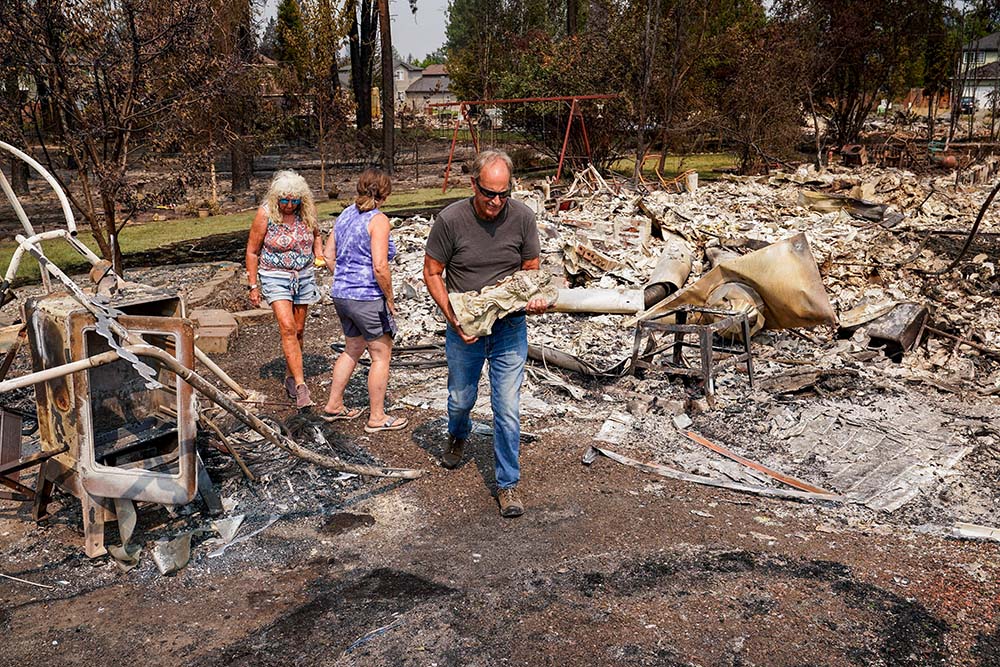 Tim Hansen carries a statue of an angel retrieved from the debris as his wife, Shelly, and Nancy Fuller survey their destroyed home in Spokane, Wash., Aug. 3, 2026, after a wildfire swept through the neighborhood. (OSV News/Reuters/David Ryder)