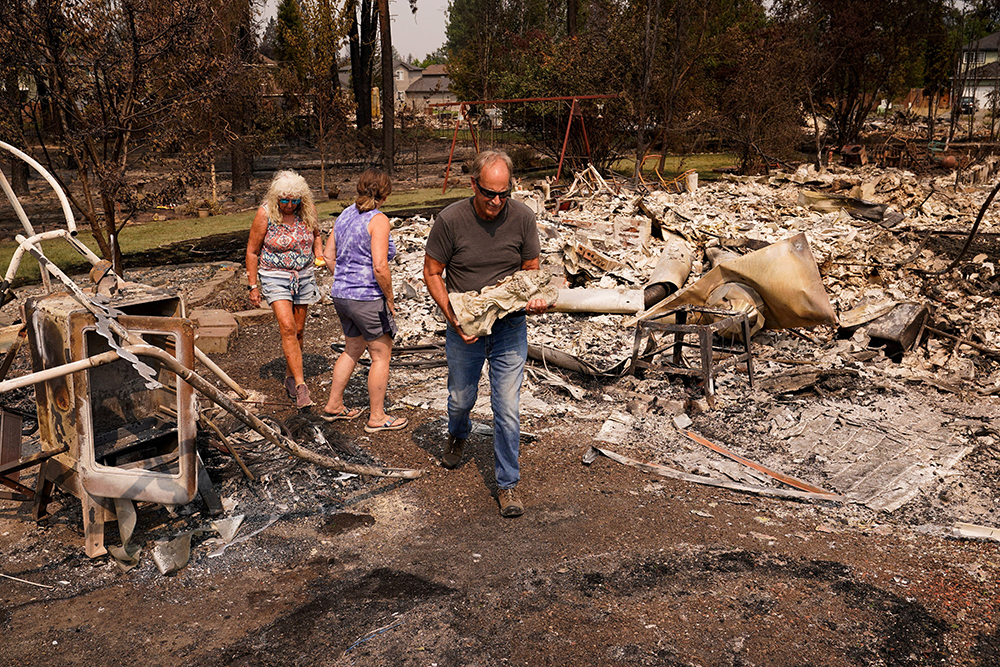 Tim Hansen carries a statue of an angel retrieved from the debris as his wife, Shelly, and Nancy Fuller survey their destroyed home in Spokane, Wash., Aug. 3, 2026, after a wildfire swept through the neighborhood. (OSV News/Reuters/David Ryder)
