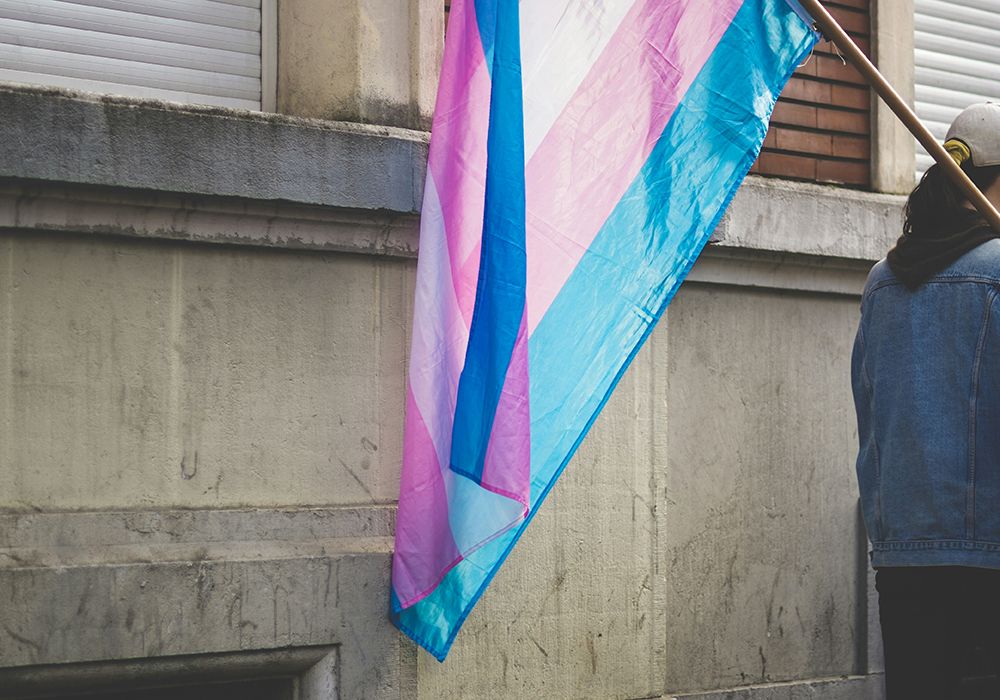 A person holds a pink, blue and white-striped flag in support of people who are transgender, standing outside near the wall of a building. (Unsplash/ev)