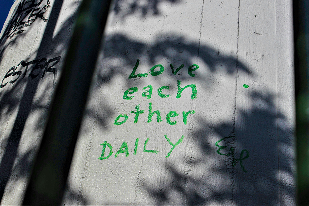 "Love each other daily," written on a fence with shadows of trees (Unsplash/Josefina Bartkova)