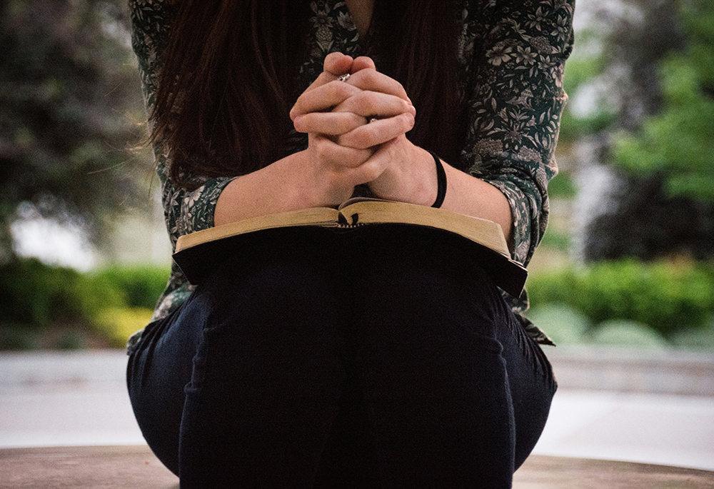 (Unsplash/Olivia Snow) A woman sits with her hands folded on top of what looks to be a Bible on her lap in her outdoor setting. Her face is not visible. (Unsplash/Olivia Snow)