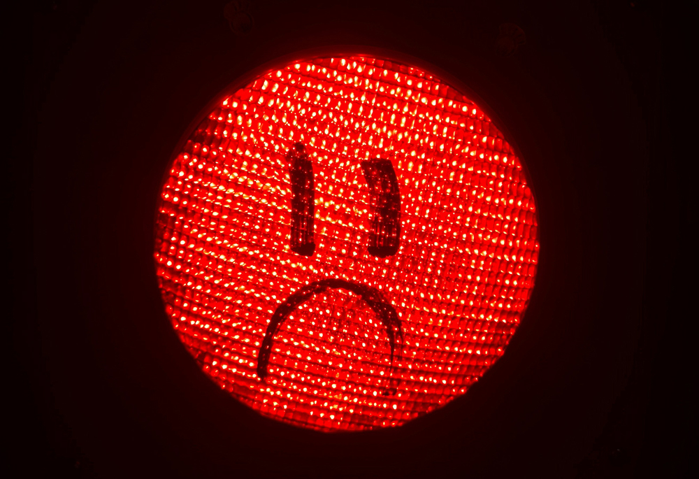 A photo illustration shows a circular red light with a drawing of a sad face on it, against a dark black background. (Unsplash/Filipe Nobre)