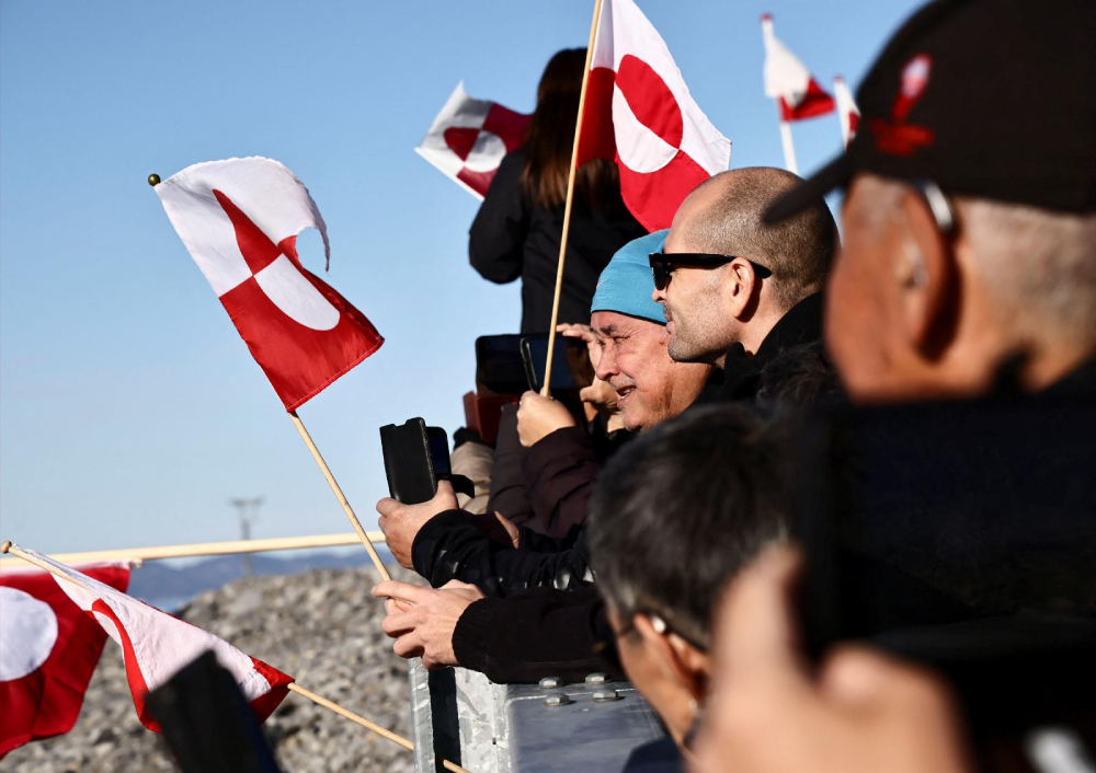 A side view of a row of people holding flags as they wait for the arrival of the Greenlandic delegation