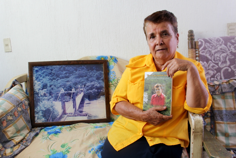 Guadalupe Lara in her home in Guadalajara, Mexico, with a copy of her memoir. A framed photograph of the historic Arcediano Bridge, which was destroyed during dam construction, rests at her side. (Tracy L. Barnett)