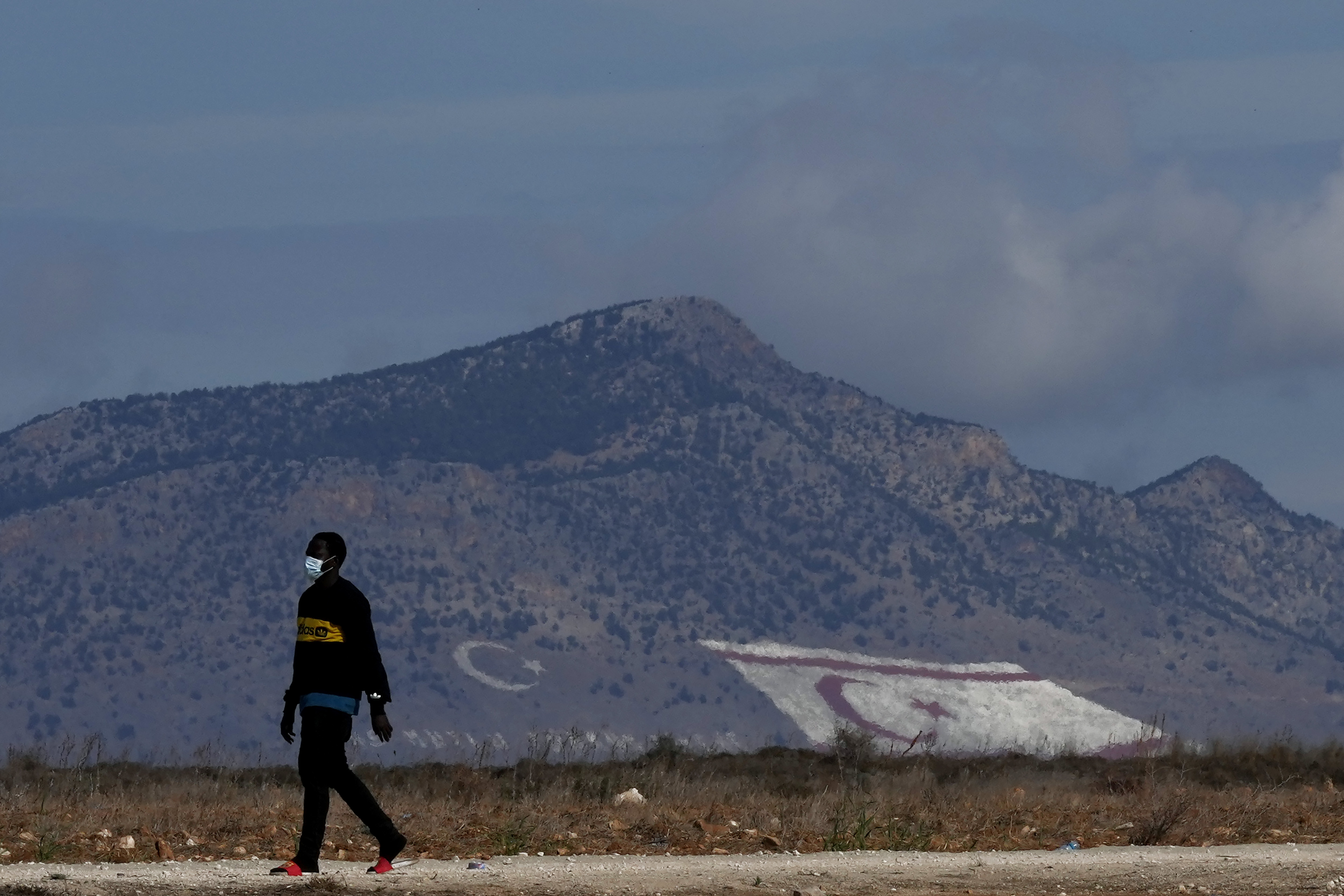 A migrant walks near Pournara migrant reception center in Kokkinotrimithia, as a giant Turkish Cypriot breakaway flag, right, and Turkish flags painted on a mountain are seen in the background in the Turkish occupied area of the ethnically divided island A migrant walks near Pournara migrant reception center in Kokkinotrimithia, as a giant Turkish Cypriot breakaway flag, right, and Turkish flags painted on a mountain are seen in the background in the Turkish occupied area of the ethnically divided island
