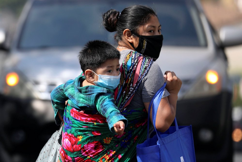 A woman in a mask carries a child in a mask on her back while toting a bag of groceries in Los Angeles May 9. (CNS/Reuters/Patrick T. Fallon)