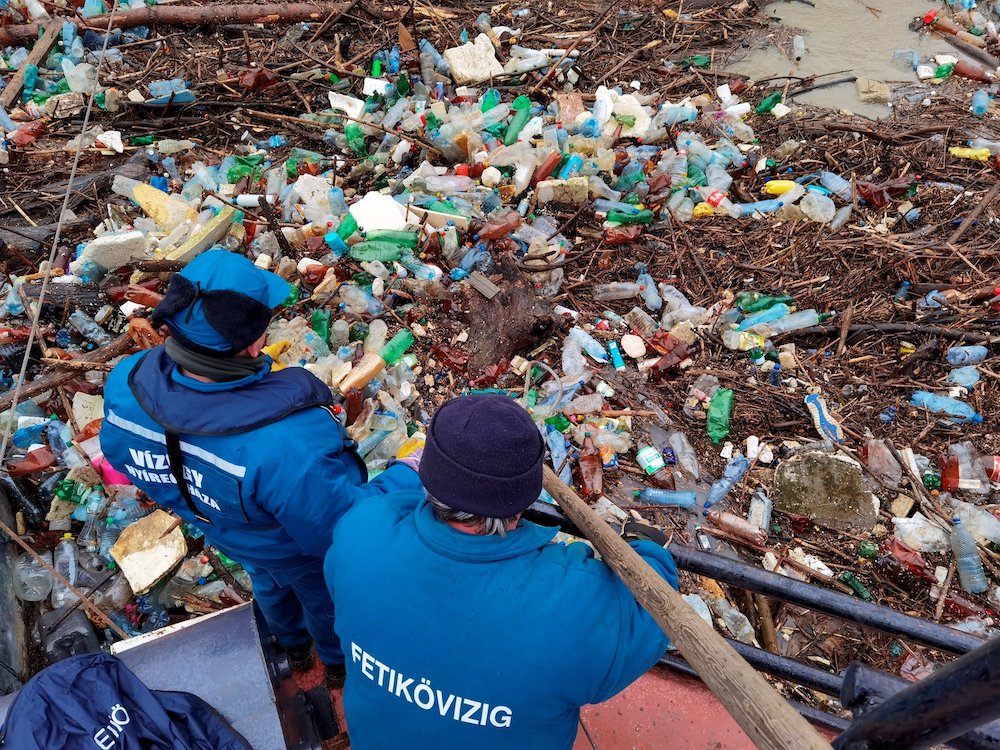 Workers collect plastic waste from the Tisza River in Vasarosnameny, Hungary, Feb. 5, 2021, after flooding. (CNS/Reuters/Hungarian Water Authority handout) Backs of two people in a boat facing floating plastic bottles and sticks in a river