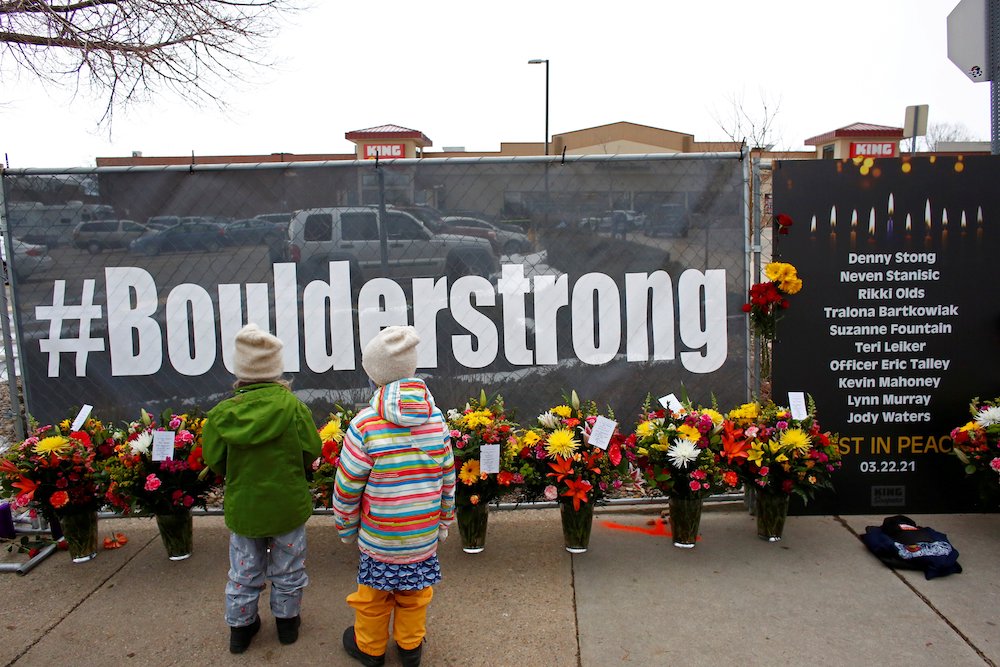 Banner on fence reads "#BolderStrong" with memorial flowers; King Sooper grocery store in background