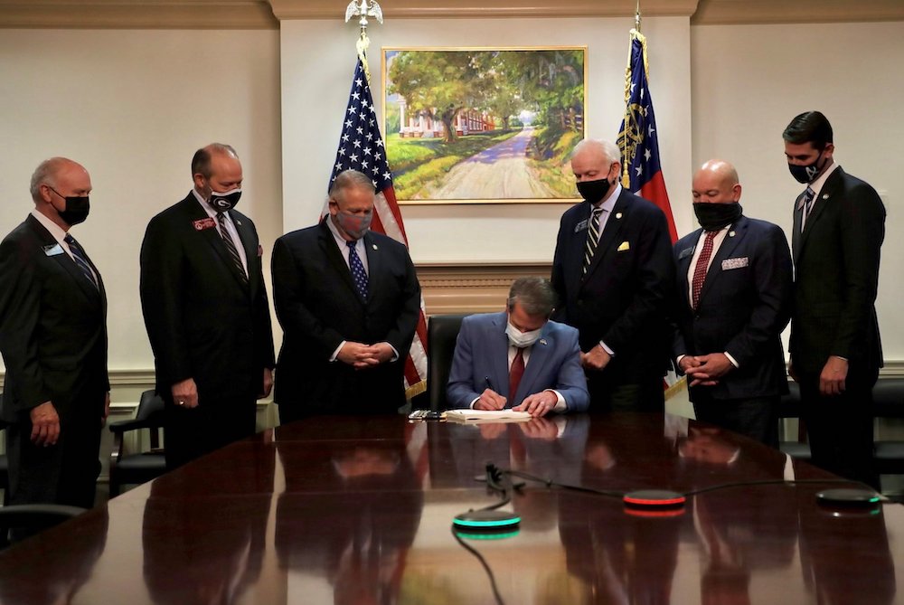Men standing around a table where governor is seated signing a bill, under a painting of a slave plantation