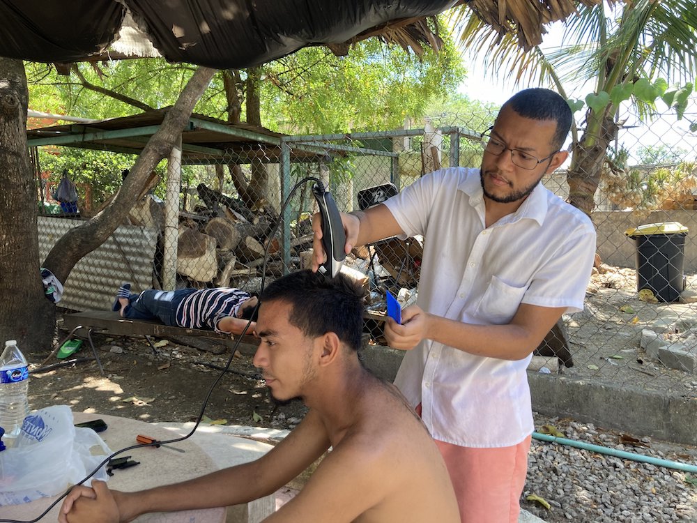 Outdoors a man stands over another seated trimming his hair with clippers