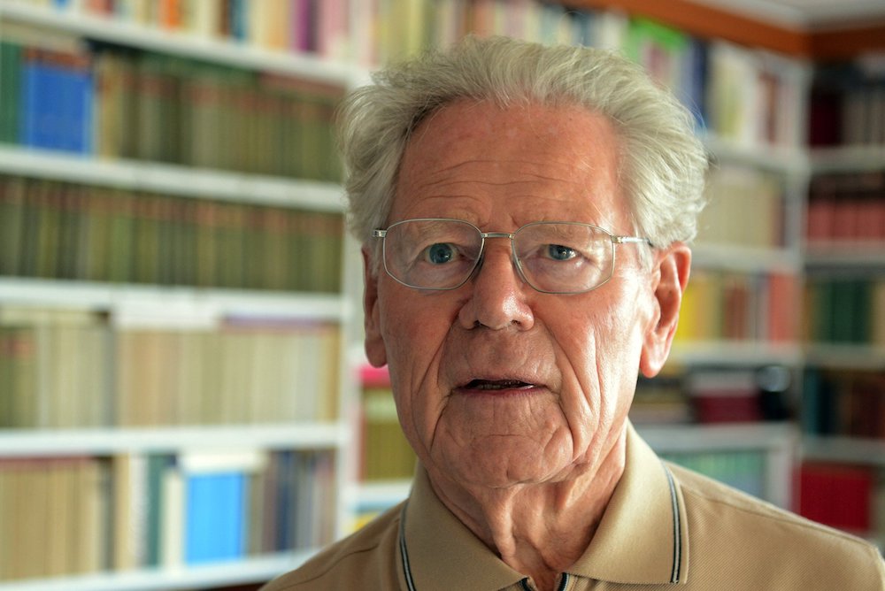 White haired man in glasses in front of books on a shelf