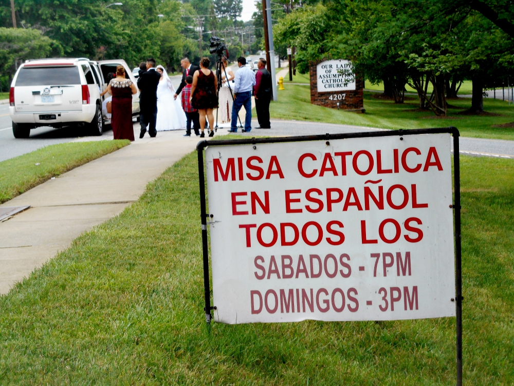 A sign announcing Masses in Spanish is seen as a wedding party gathers at Our Lady of the Assumption Church in Charlotte, North Carolina. (NCR photo/Peter Feuerherd)