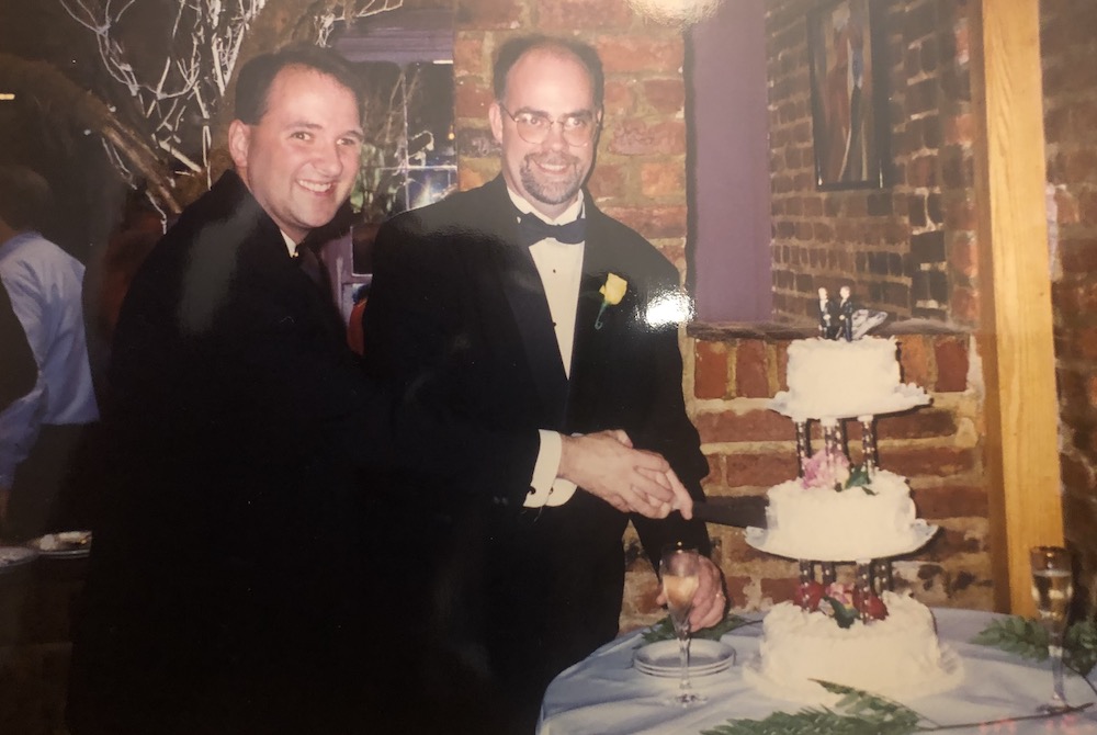 Two men look at the camera holding hands at a table with a wedding cake