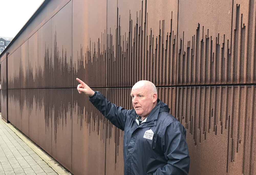 Paul Doherty stands in front of one of the buildings hit by the soldiers' bullets, which is now adorned with an artistic visualization of the sound of the civil rights anthem "We Shall Overcome." (NCR photo/Claude Colart)