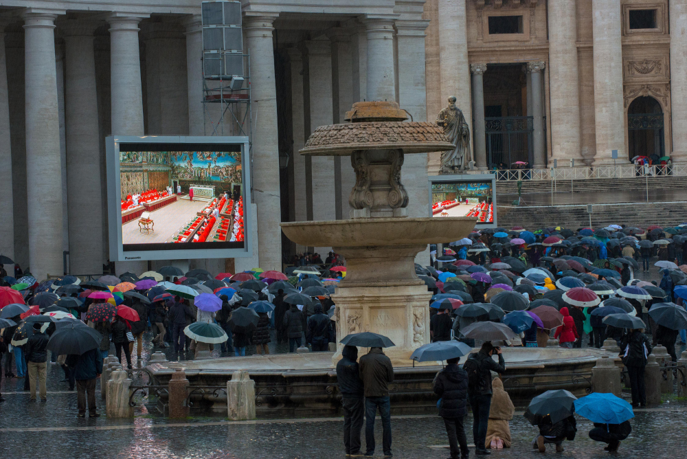 Faithful visitors holding umbrellas look at a giant screen showing the start of the 2013 papal conclave, under a statue of St. Peter on St. Peter's Square at the Vatican. (RNS photo/Andrea Sabbadini)