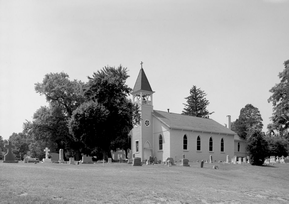 Black and white image of a small church with bell tower in field with trees and grave markers