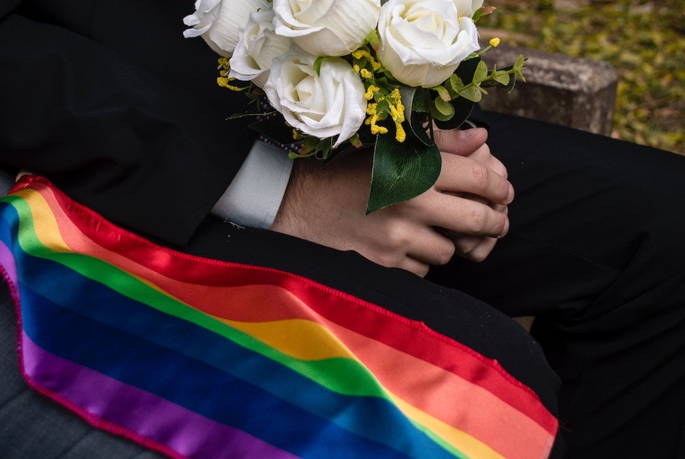Rainbow flag next to seated man in black suit holding a bouquet of white roses