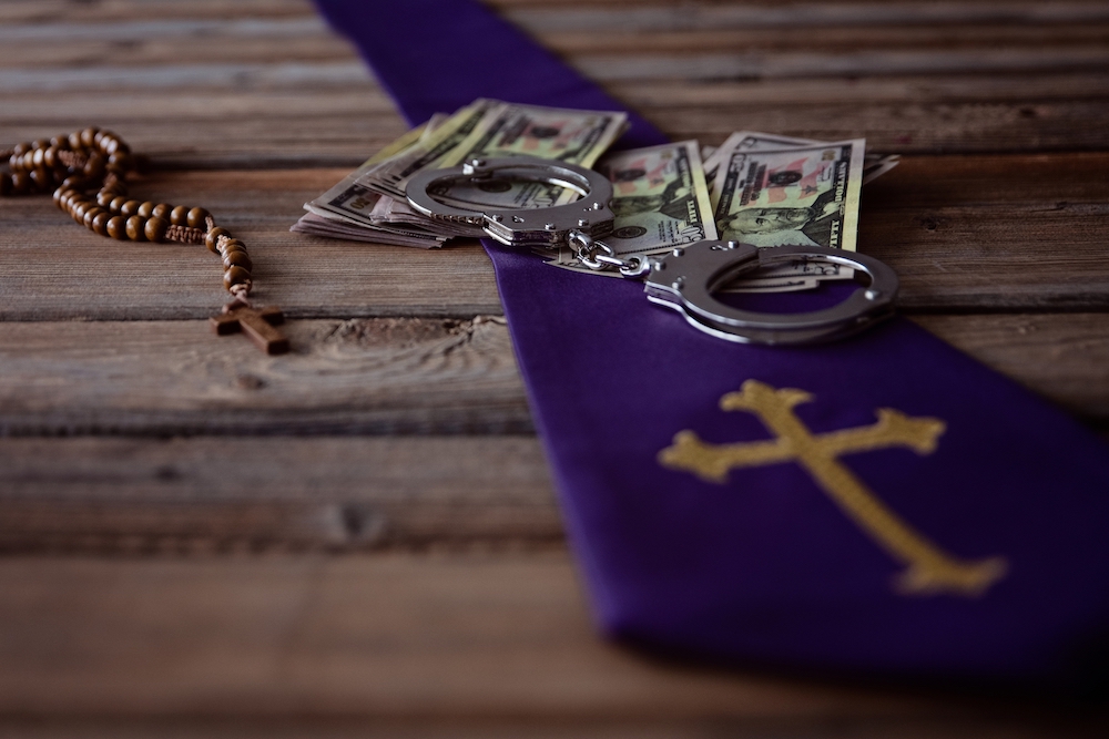 Image of a rosary, US currency, handcuffs and a cross on a wooden table (Dreamstime/Djedzura)