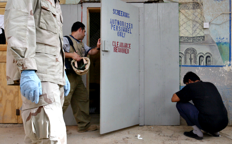 A private contractor involved in prisoner interrogation enters an interrogation room as an Iraqi detainee squats outside in a processing center at the Abu Ghraib Prison on the outskirts of Baghdad in May 2004. (AP Photo/John Moore)