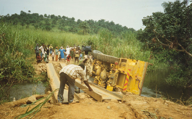 A caravan of School Sisters of Notre Dame, schoolgirls and others fleeing rebel violence in November 1992 in Sierra Leone waits on a back road as locals repair a bridge that buckled, overturning the caravan’s truck.