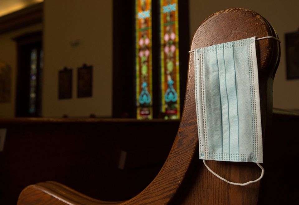 In this illustration photo, a disposable medical mask hangs on the side of a church pew. (CNS/ The Catholic Spirit/Dave Hrbacek)