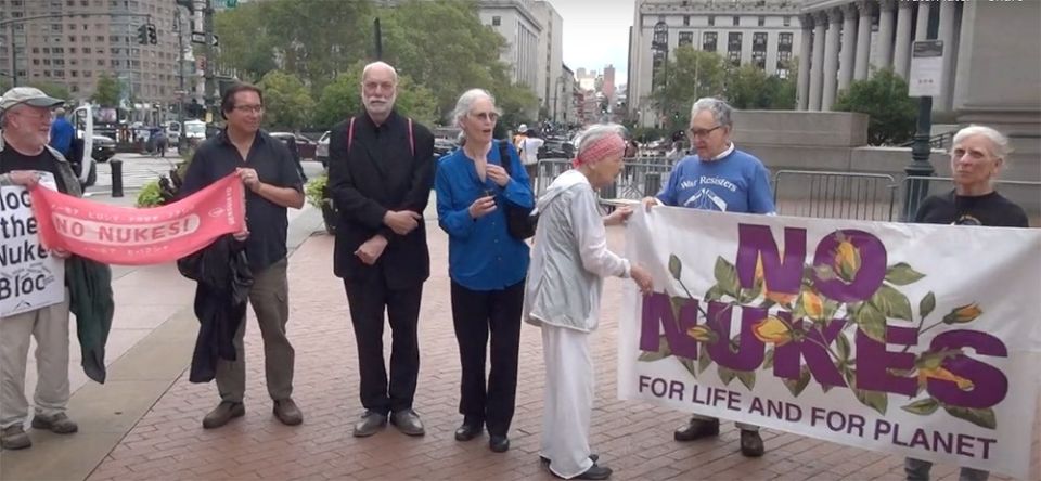A group of older adults stand outside the courthouse with signs with messages like "No Nukes for life and for planet"