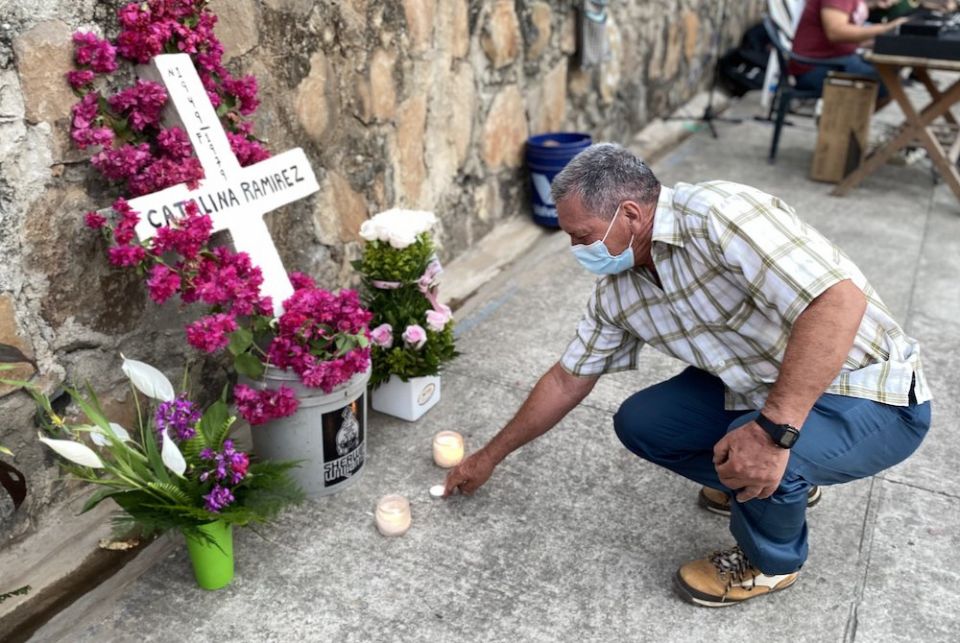A man in a mask kneeling to light a candle before a wooden cross in a bucket of pink flowers and bearing the name of his sister