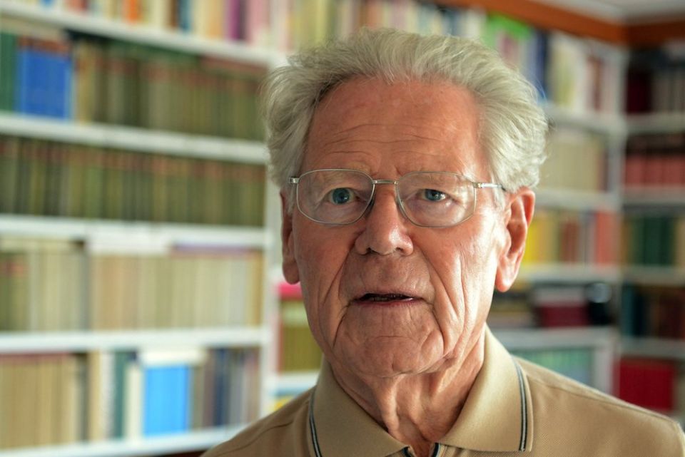 White haired man in glasses in front of books on a shelf