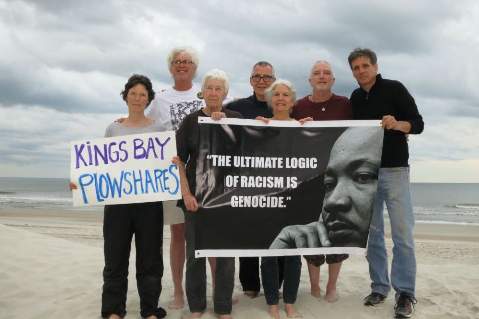 Seven people on a beach holding signs; one says "Kings Bay Plowshares," the other features a portrait of Martin Luther King, Jr.