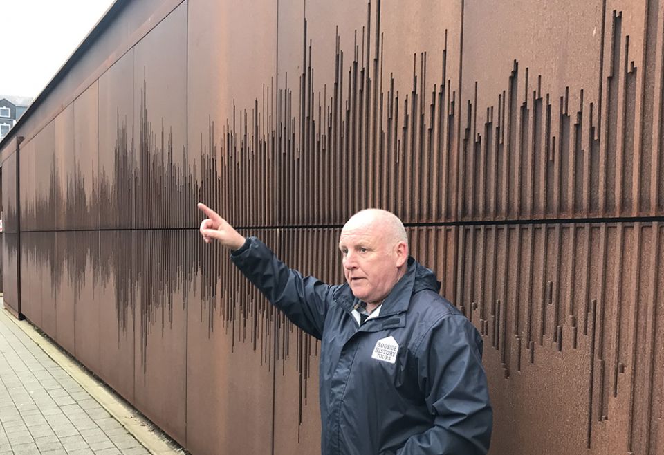 Paul Doherty stands in front of one of the buildings hit by the soldiers' bullets, which is now adorned with an artistic visualization of the sound of the civil rights anthem "We Shall Overcome." (NCR photo/Claude Colart) Paul Doherty stands in front of one of the buildings hit by the soldiers' bullets, which is now adorned with an artistic visualization of the sound of the civil rights anthem "We Shall Overcome." (NCR photo/Claude Colart)