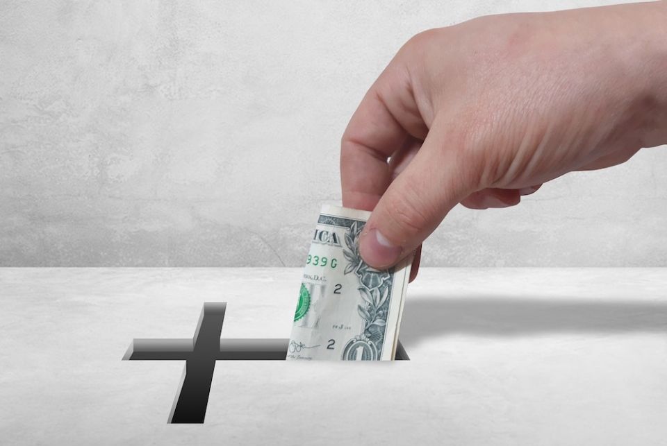 A parish in Diocese of Rockville Centre is recovering from high expenses under its former pastor, Fr. Charles Mangano, who was removed in mid-2018. And some parishioners say that there has been retribution for their questioning. (Pixabay/JeffJacobs1990) Hand putting or taking a folded dollar bill into a cross-shaped slot on collection box