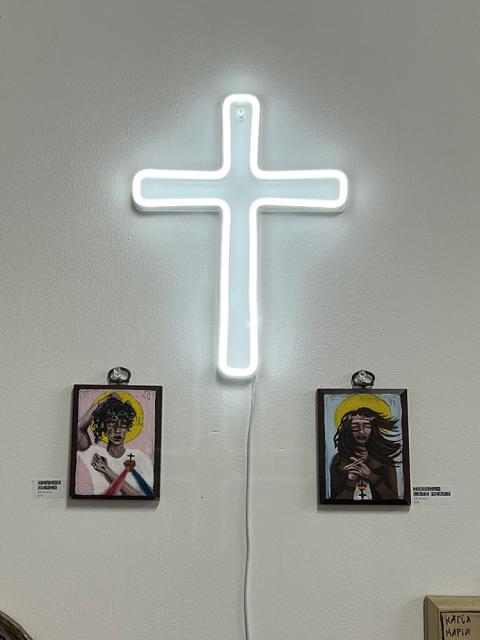 A neon cross on a white wall between two of Gracie Morbitzer's icons