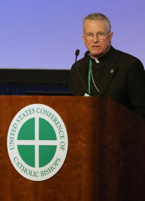 Military Services Archbishop Timothy Broglio stands at lectern with green and white USCCB logo.