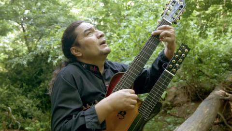 Man with long hair plays stringed instrument amidst greenery