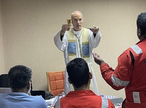 Baltimore Auxiliary Bishop Parker raises Eucharistic elements as he says Mass in ship's hull. Stranded sailors pictured from behind, facing Parker. 